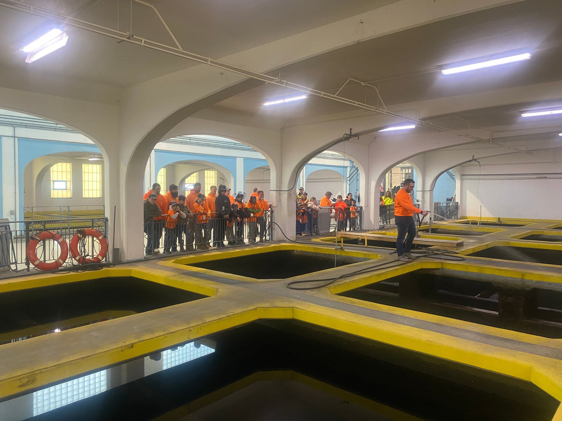 People in orange vests standing near tanks of water in a large, arched building.