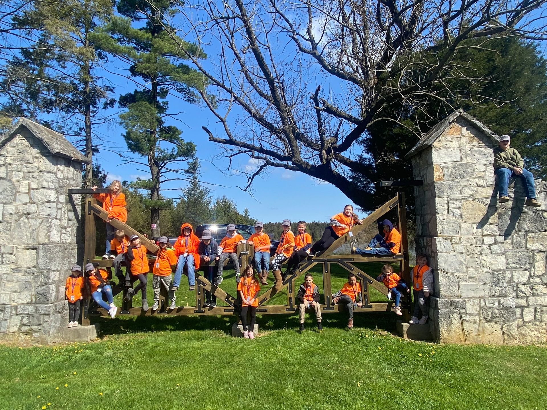 Group of people in orange vests pose on a gate in front of stone structures and trees.