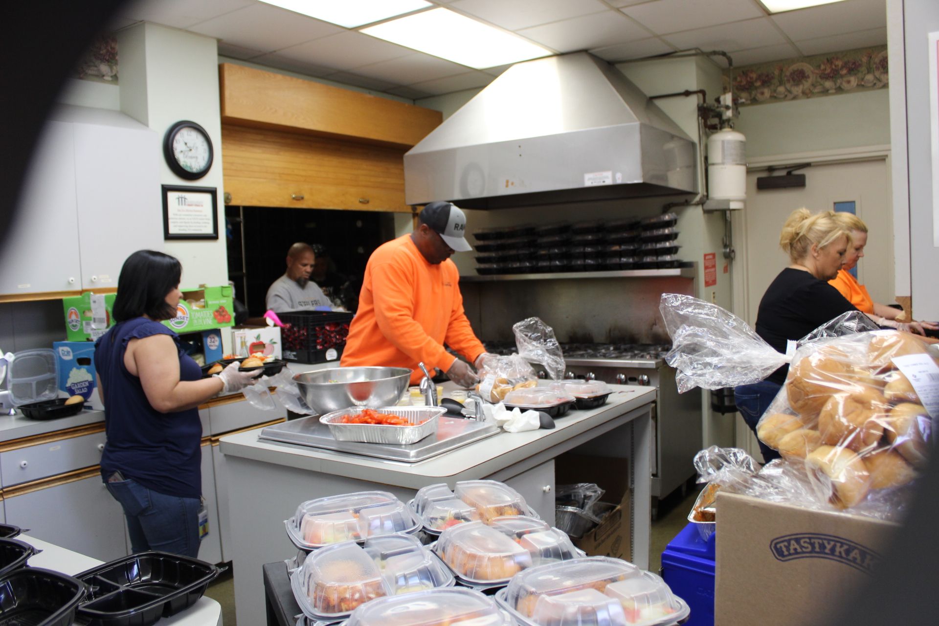 People preparing food in a kitchen. Several people are packaging meals and working near a large stove.