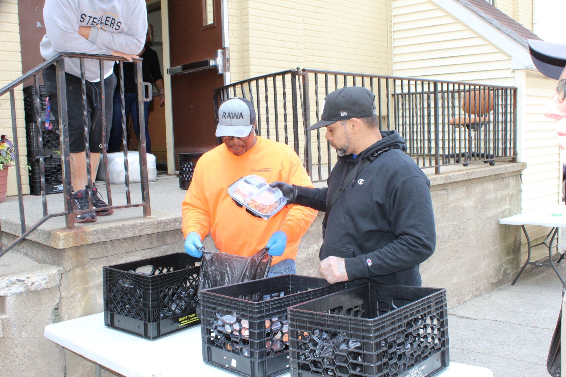 Two men sorting items into crates outside a building with steps. One man wears orange.