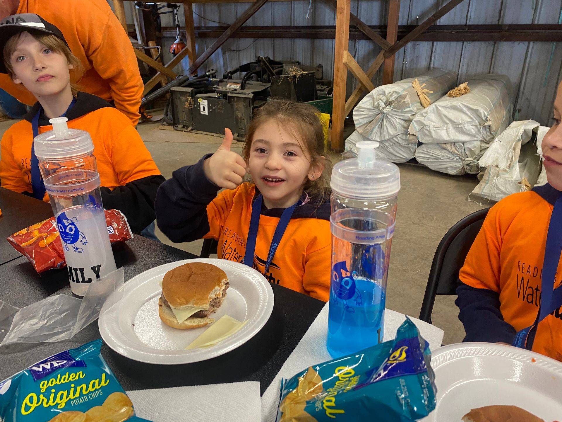 Children eating at a table, one giving a thumbs-up. They wear orange shirts and have food/drinks in a warehouse setting.