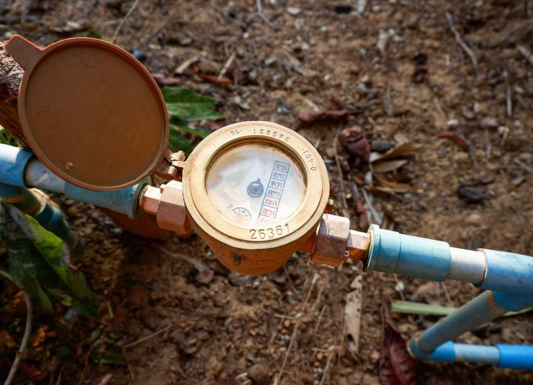 Water meter on blue pipes, brass housing with brown cover in a dirt setting.