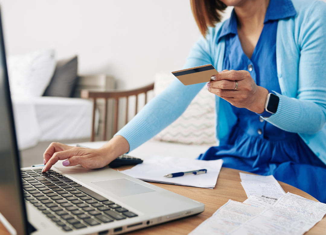 Person typing on a laptop, holding a credit card, at a desk with papers and a pen.