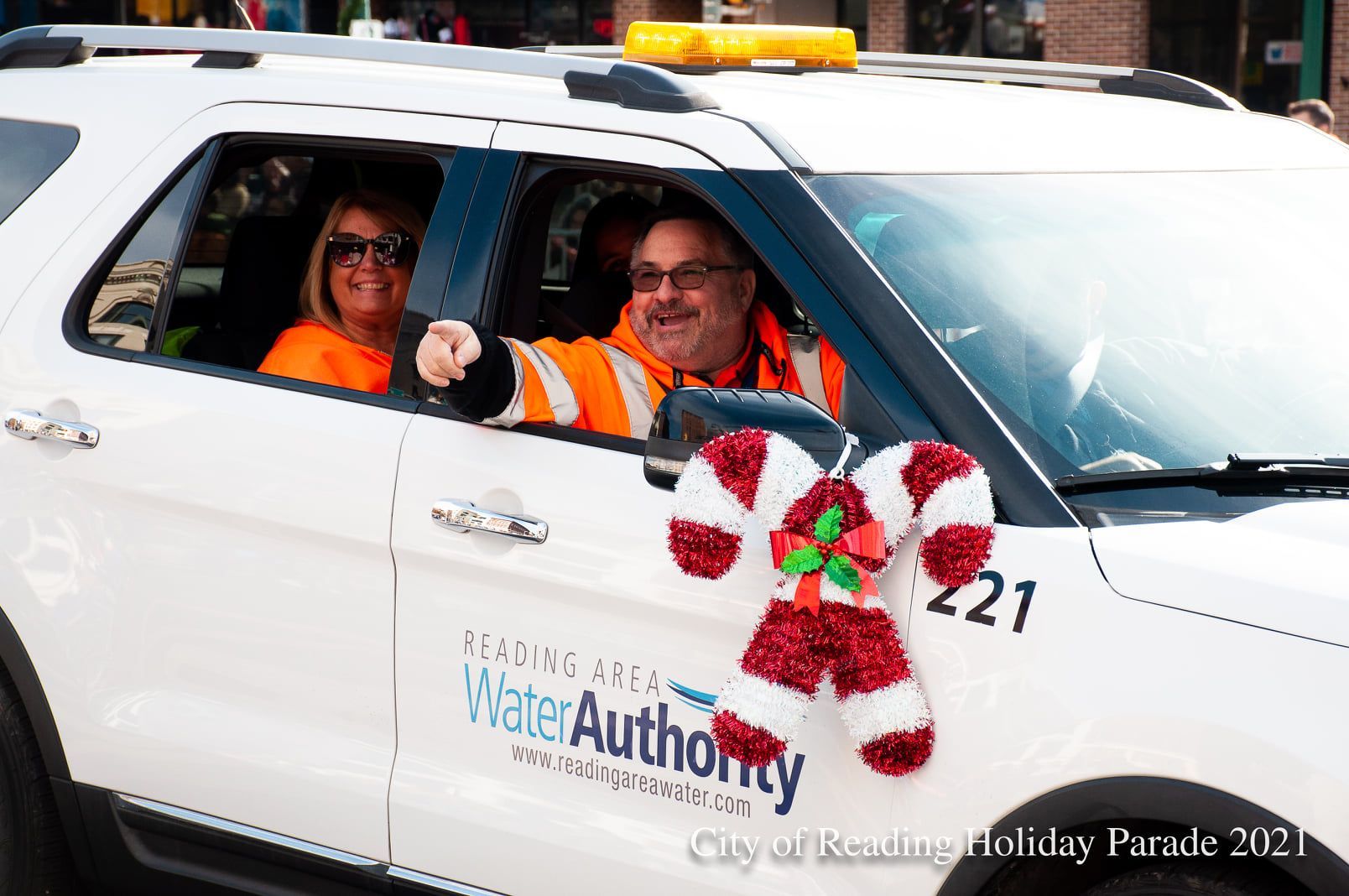 White SUV in a parade decorated with a candy cane; two people inside waving.