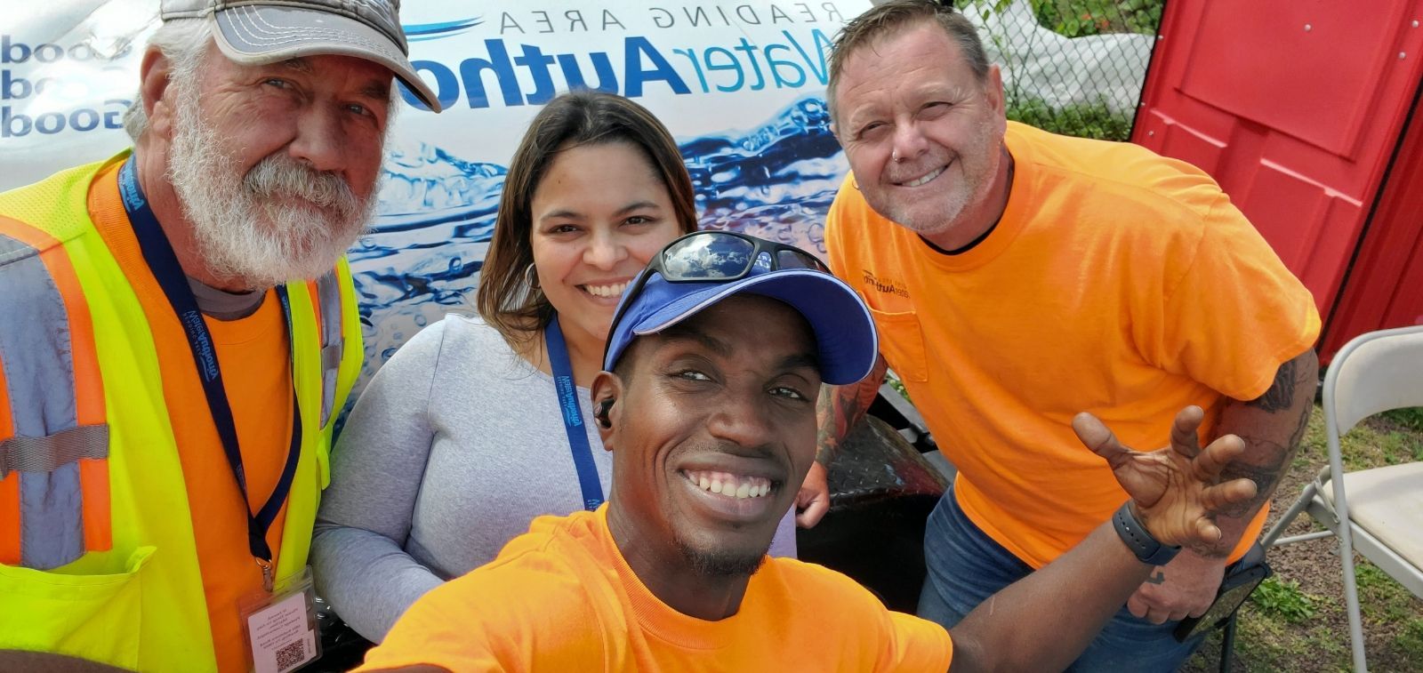 Four people smiling at the camera, posing for a selfie. They are outside near a banner that has blue water.