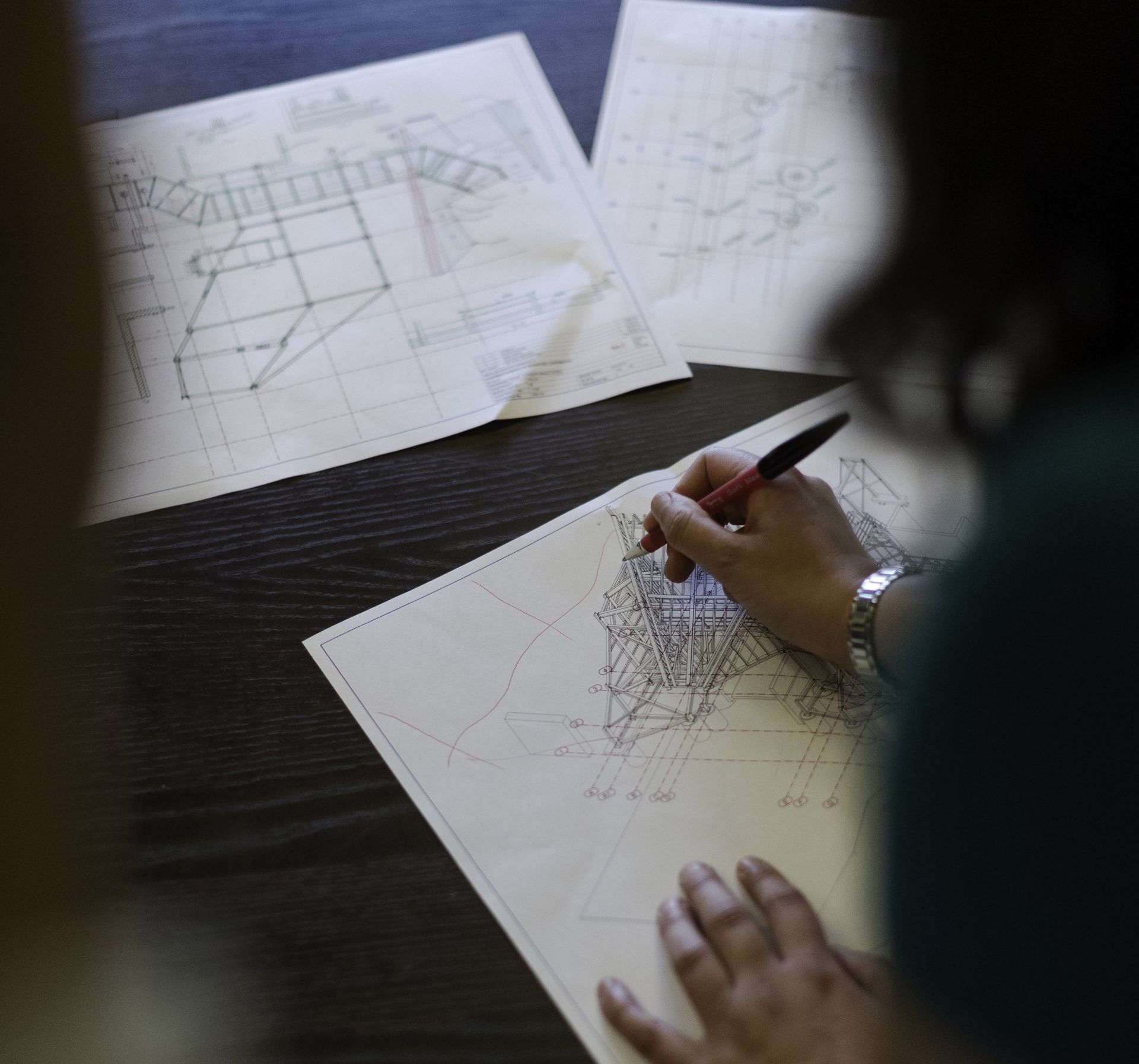 A person sketching architectural designs on a blueprint with a pen; other blueprints on the table.