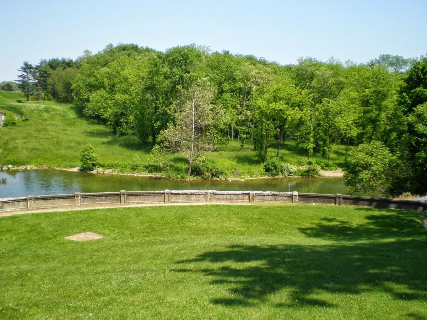Green lawn leads to a lake, with trees and a hill in the background on a sunny day.