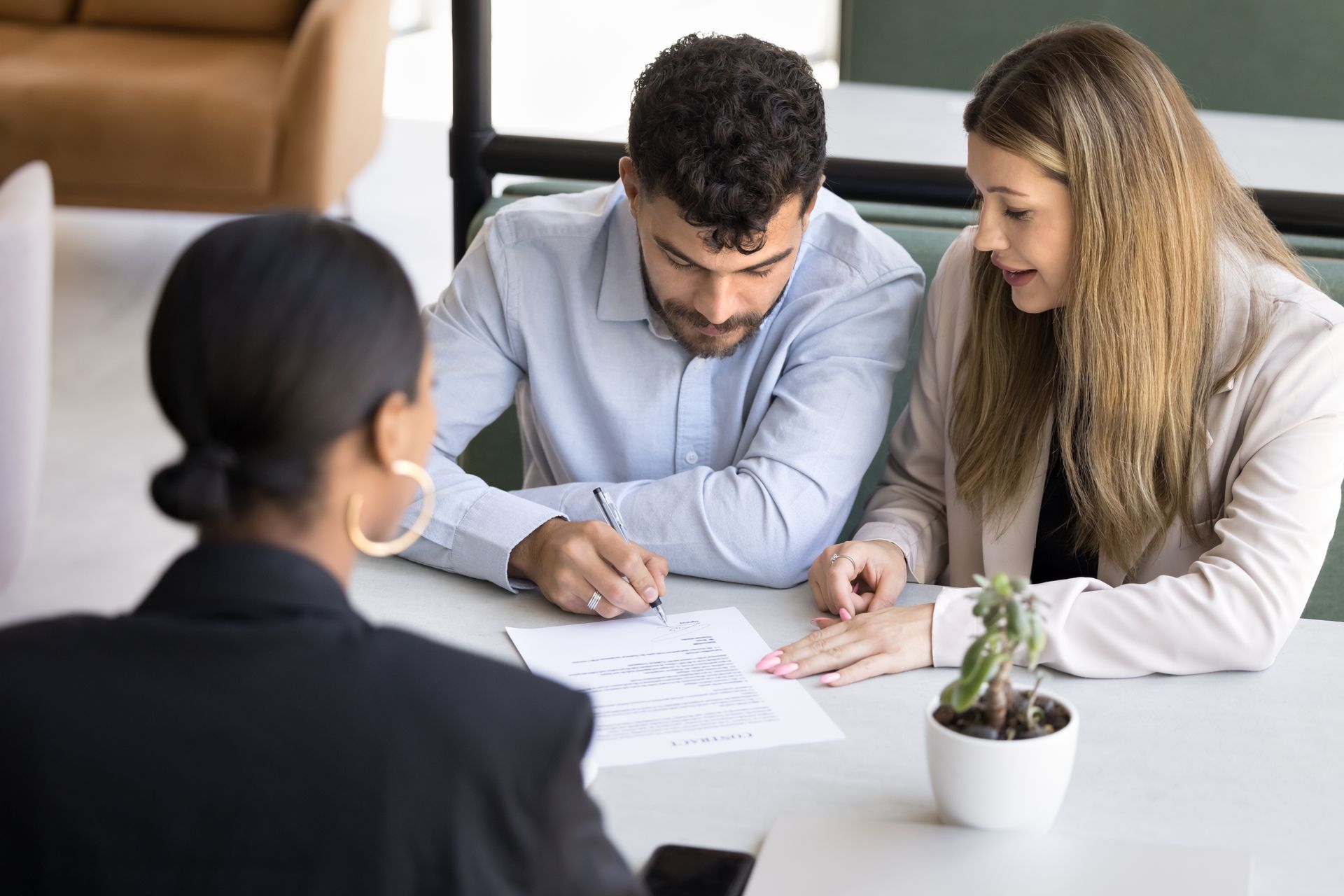 Three people reviewing documents at a table in a bright office, with a small potted plant nearby.