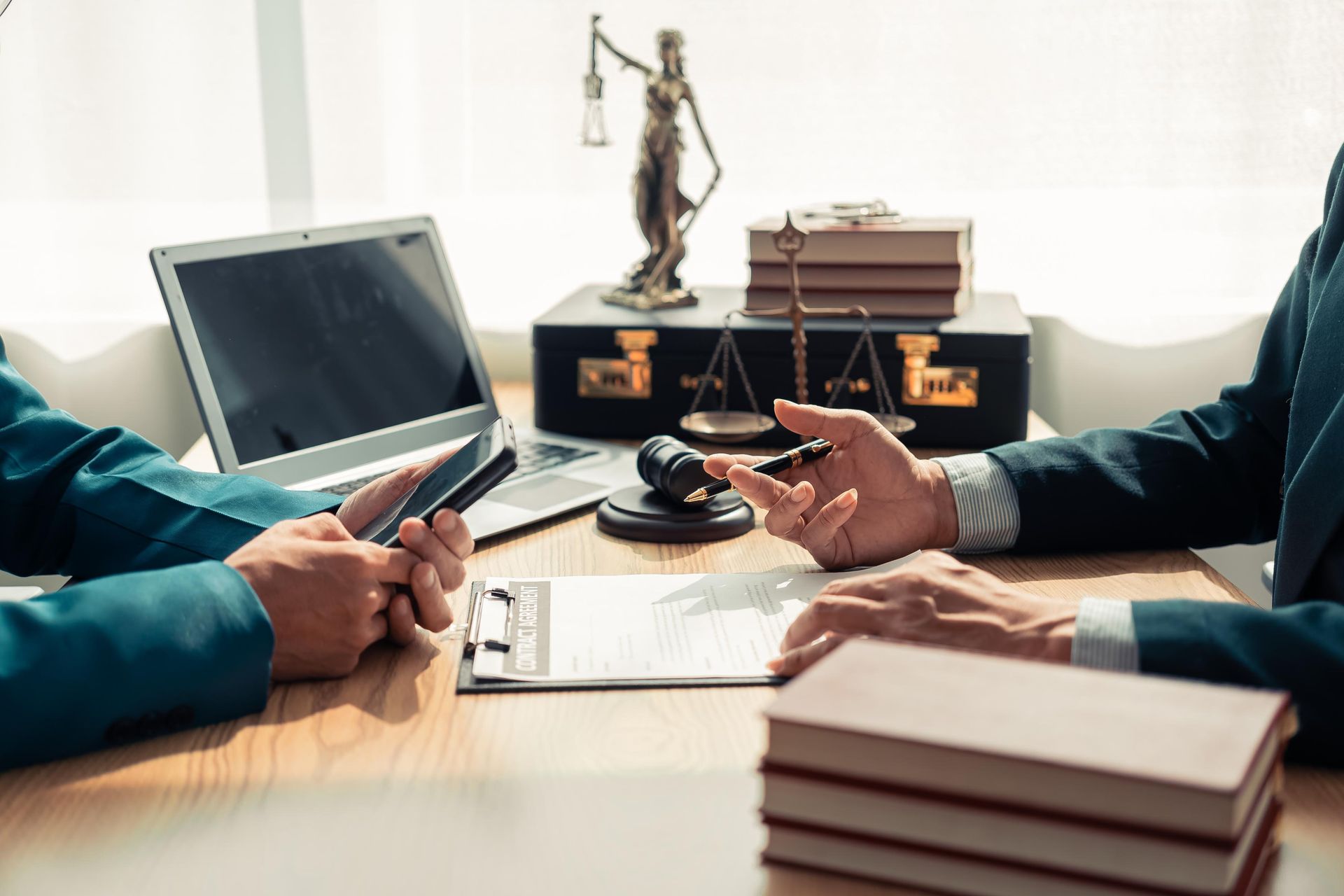 Two people discussing documents at a desk with a laptop, scales of justice, and law books in a legal office.