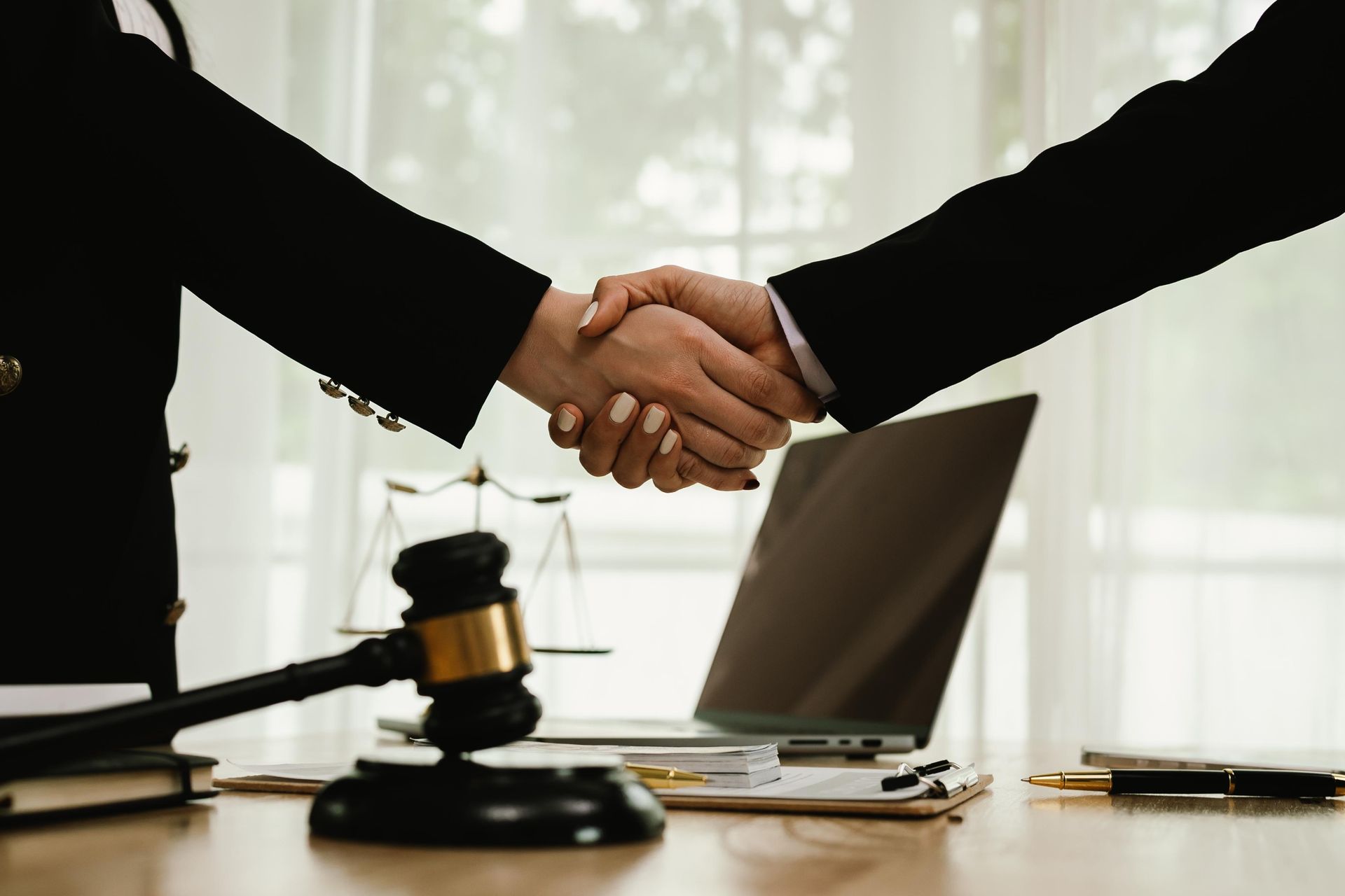 Two people in suits shaking hands across a desk with a laptop and gavel, suggesting a legal agreement