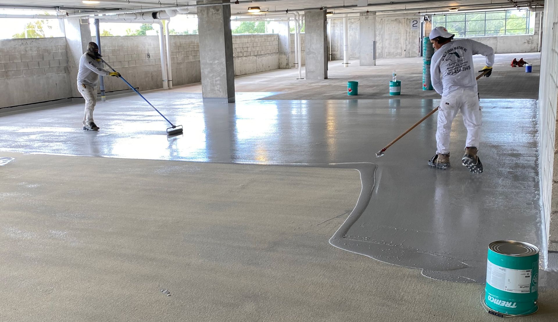 Workers applying gray coating to a concrete floor in a parking garage.