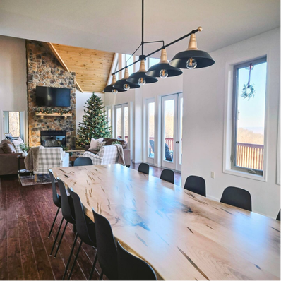 A modern dining room with a long light-wood table, black chairs, and pendant lighting, opening to a cozy living area.