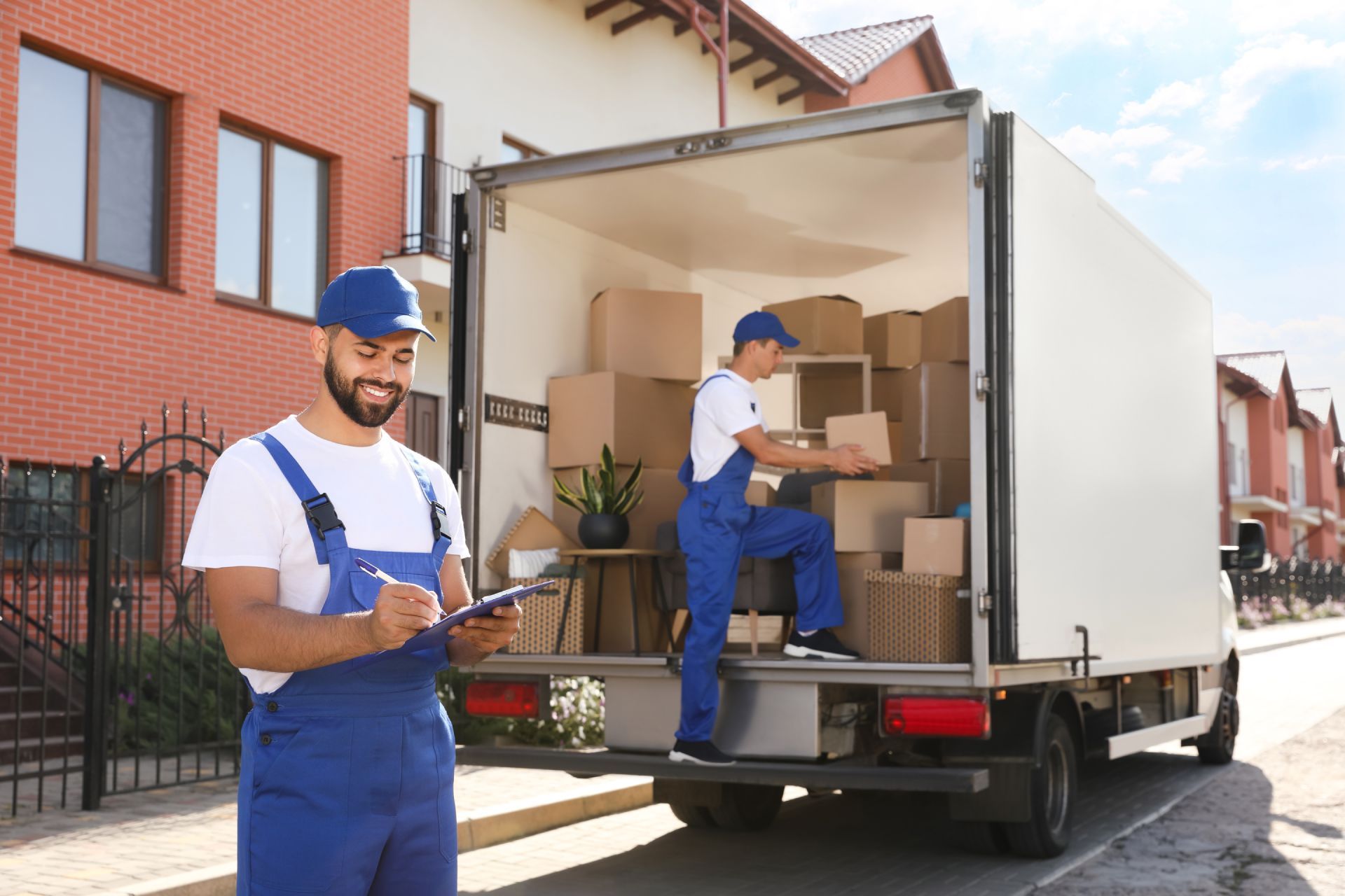 Movers loading boxes into a truck, one writing on a clipboard, blue and white uniforms, houses in the background.