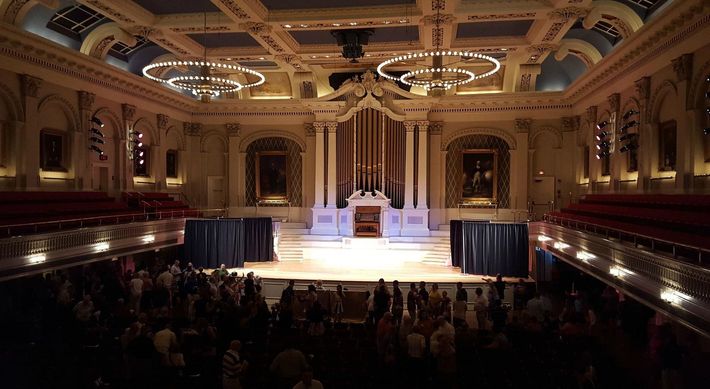 Grand ballroom with ornate ceiling, chandeliers, stage, and audience seated in the dark foreground