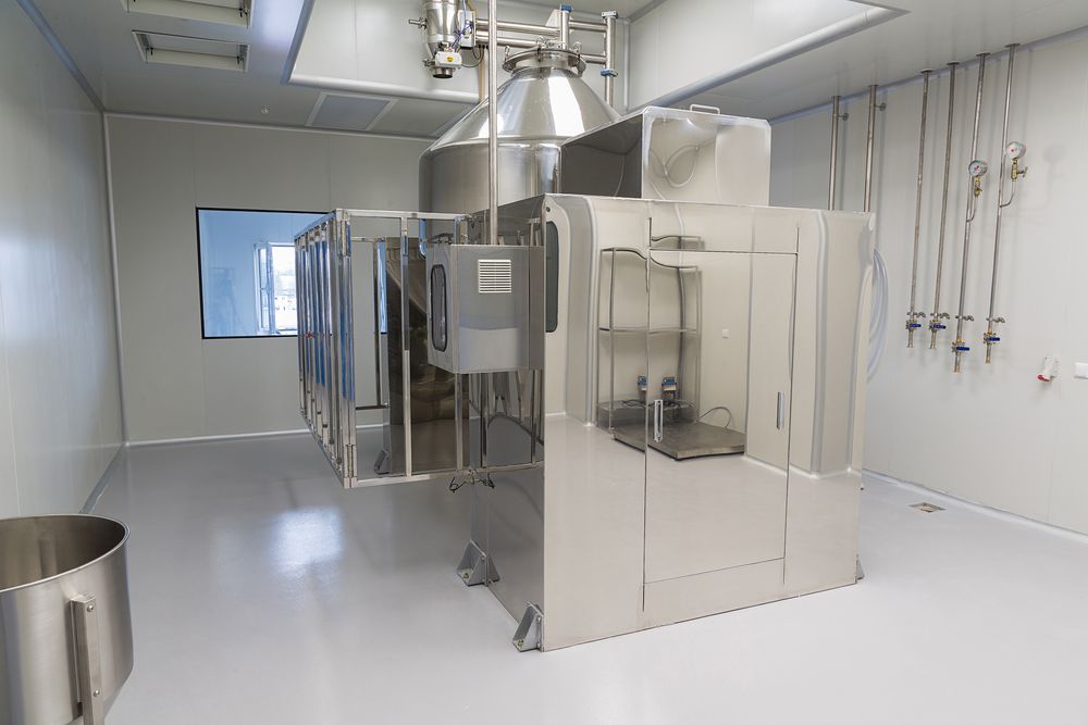 Stainless steel industrial pharmaceutical equipment in a clean room setting with white walls and a polished floor.
