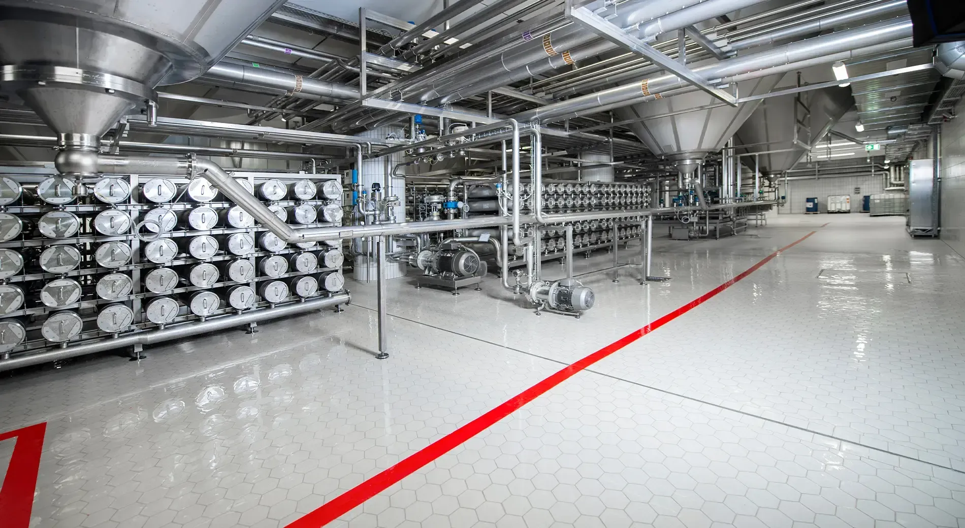 An industrial processing plant with shiny stainless steel equipment, pipes, and a bright, sterile floor with red lines.