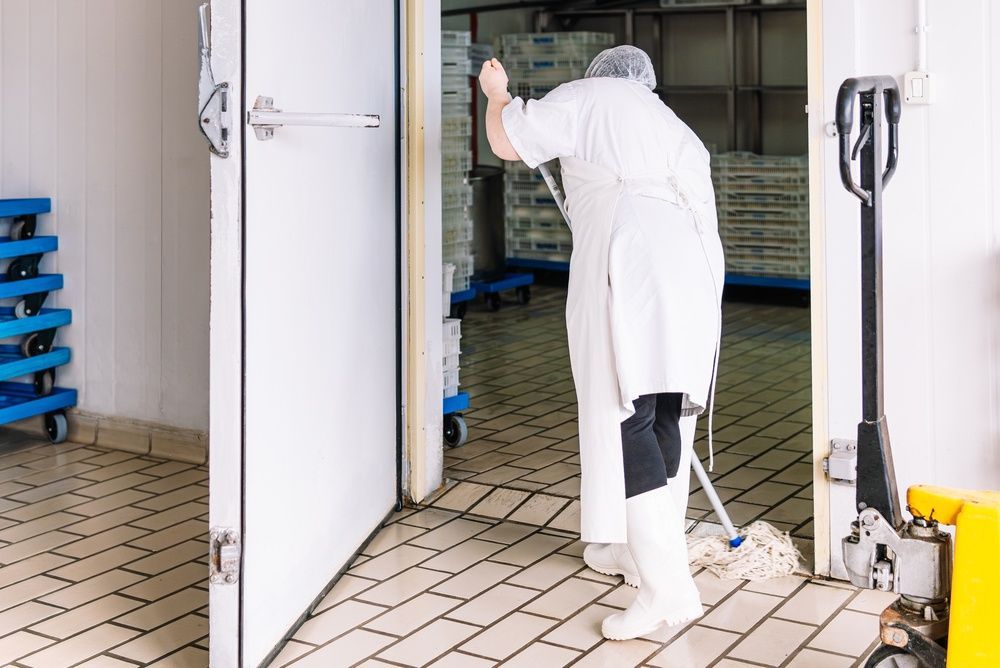 A person in white sanitary work clothing mopping the tiled floor of a walk-in industrial freezer.