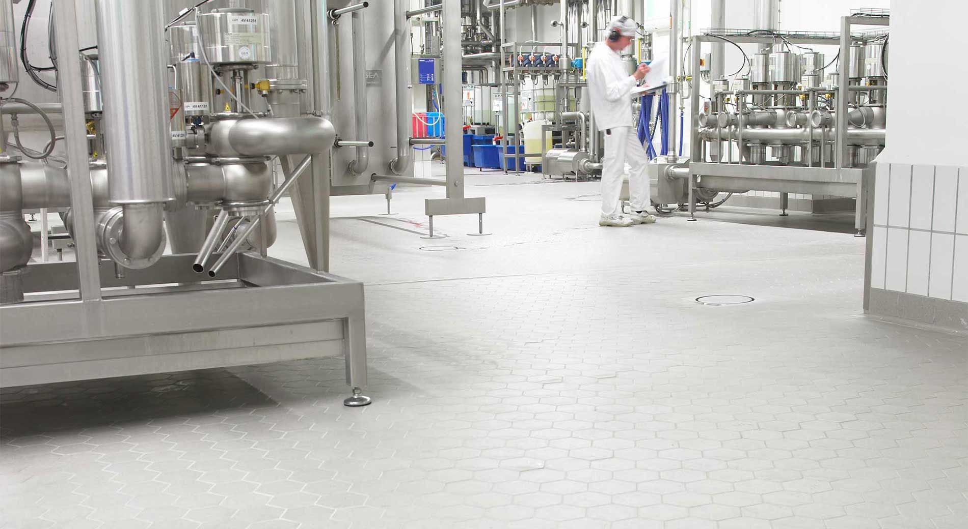 A person in white protective work clothing stands in a bright, sterile industrial facility inspecting stainless steel pipes.