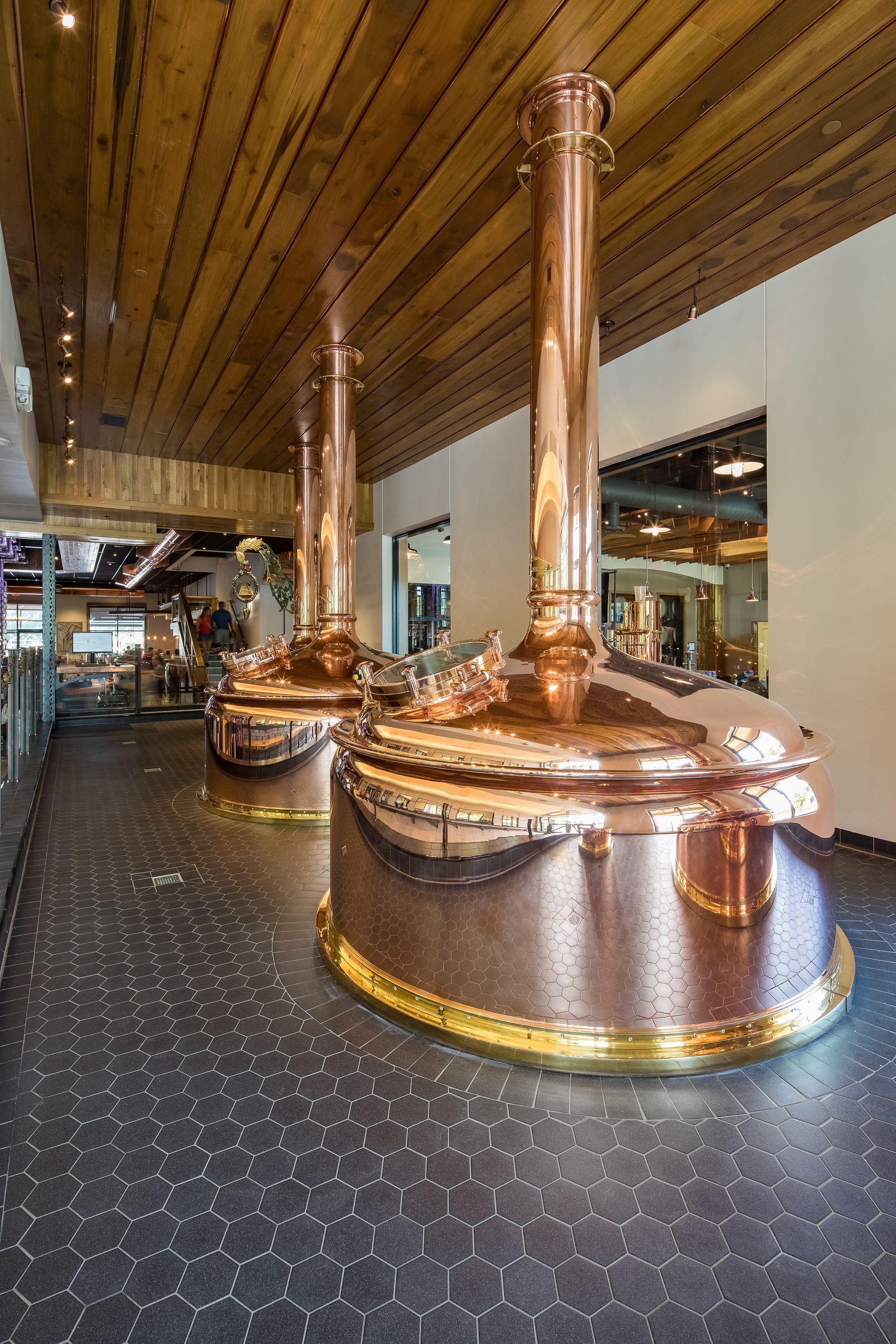 Two large, polished copper brewing kettles with vertical chimneys stand in a brewery with wooden ceilings and dark floors.