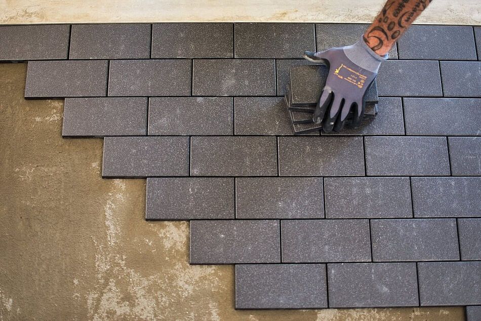 A gloved hand places a gray rectangular paving stone onto a sand base, continuing a staggered masonry pattern.