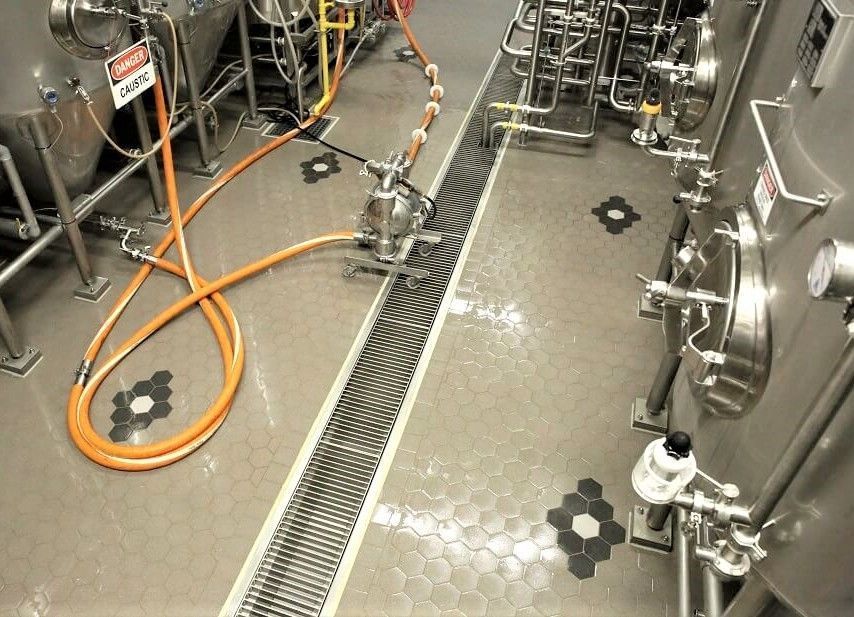 Industrial floor with a central metal drainage grate, surrounded by stainless steel tanks and orange hoses.