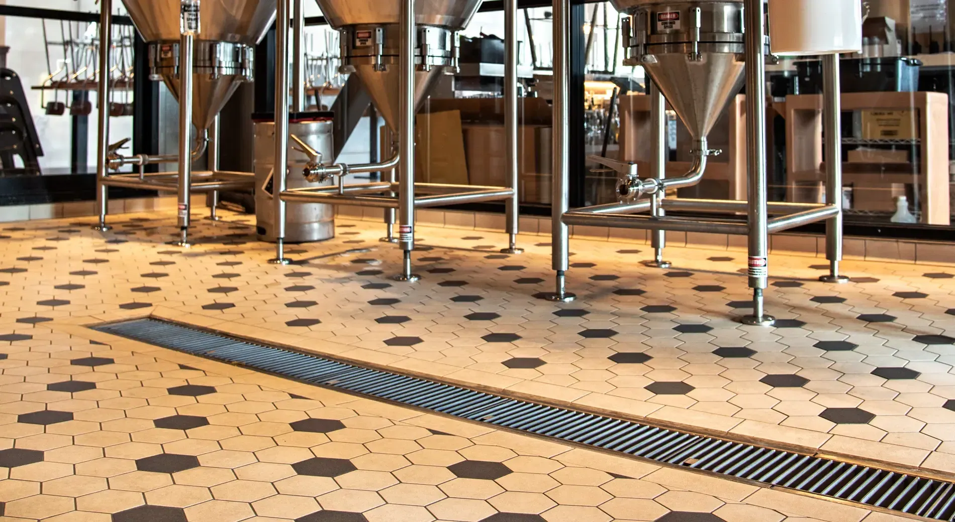 Industrial brewery room with stainless steel fermentation tanks above a tiled floor and a metal drainage grate.