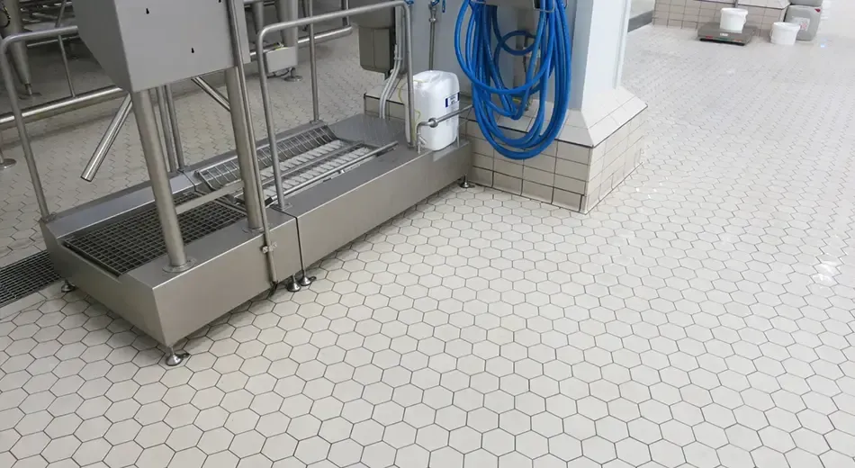 A stainless steel industrial boot cleaner station stands on a light-colored hexagonal tiled floor.