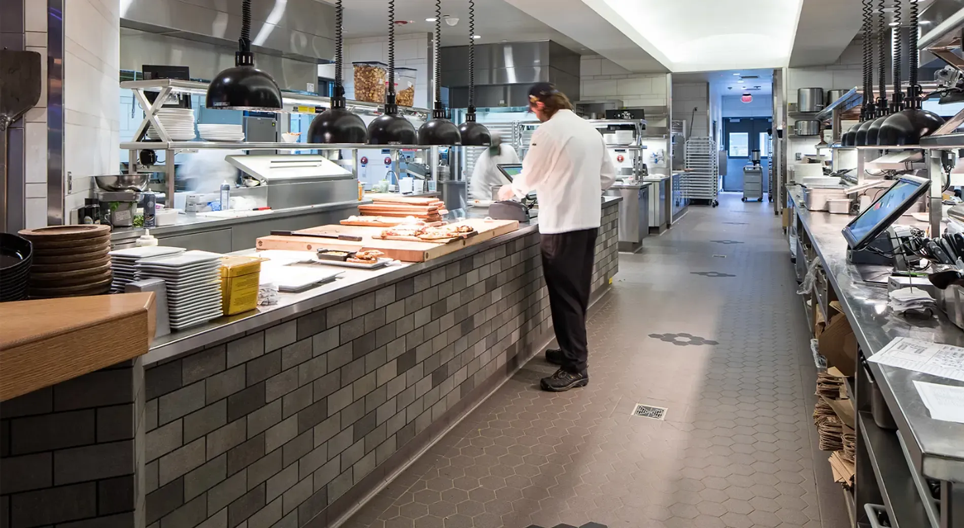 A chef in a white coat stands at a long, brick-fronted kitchen prep counter in a busy restaurant kitchen.