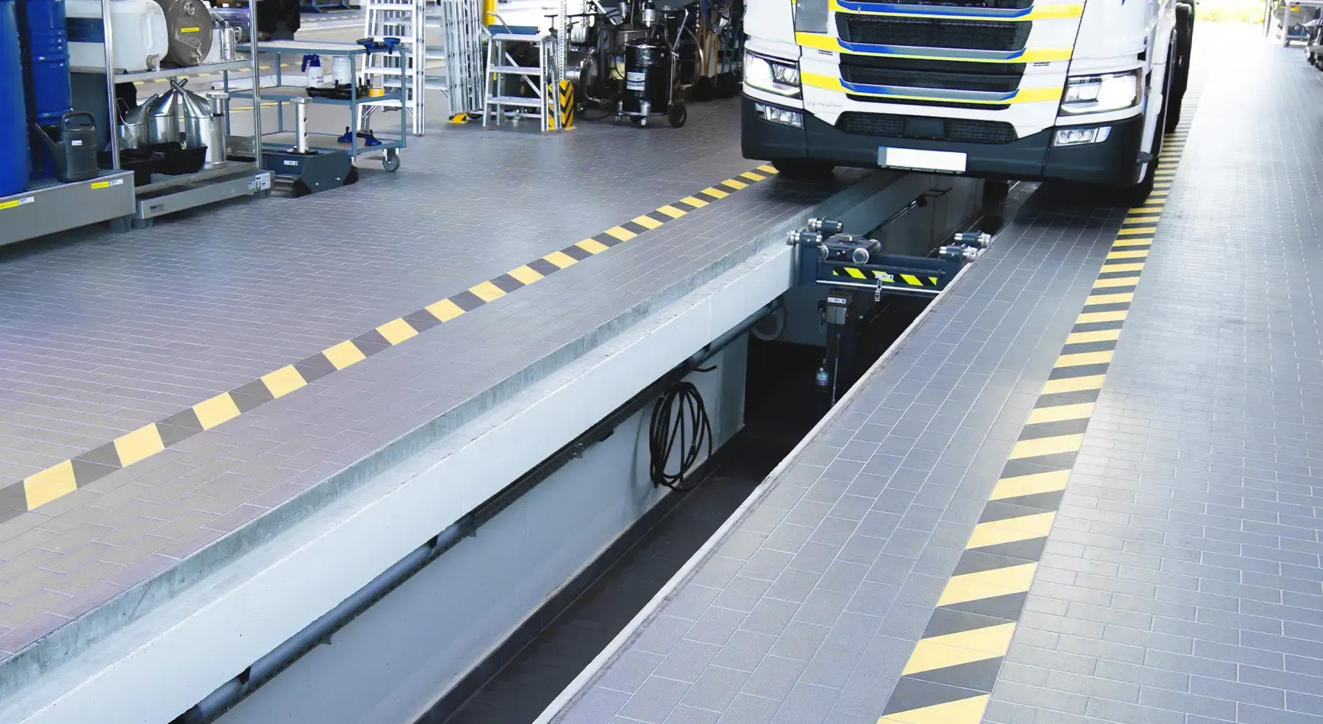 A truck parked over a service inspection pit in a workshop, with yellow and black safety markings on the floor.
