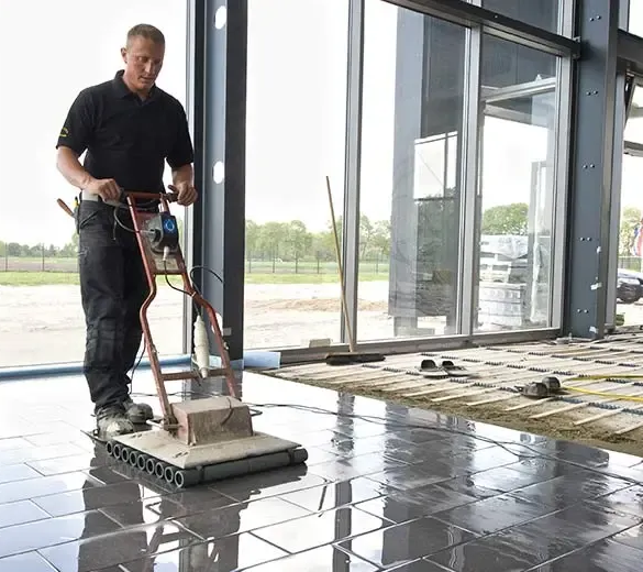 Worker operating a floor-cleaning machine on a glossy tiled indoor floor near large windows