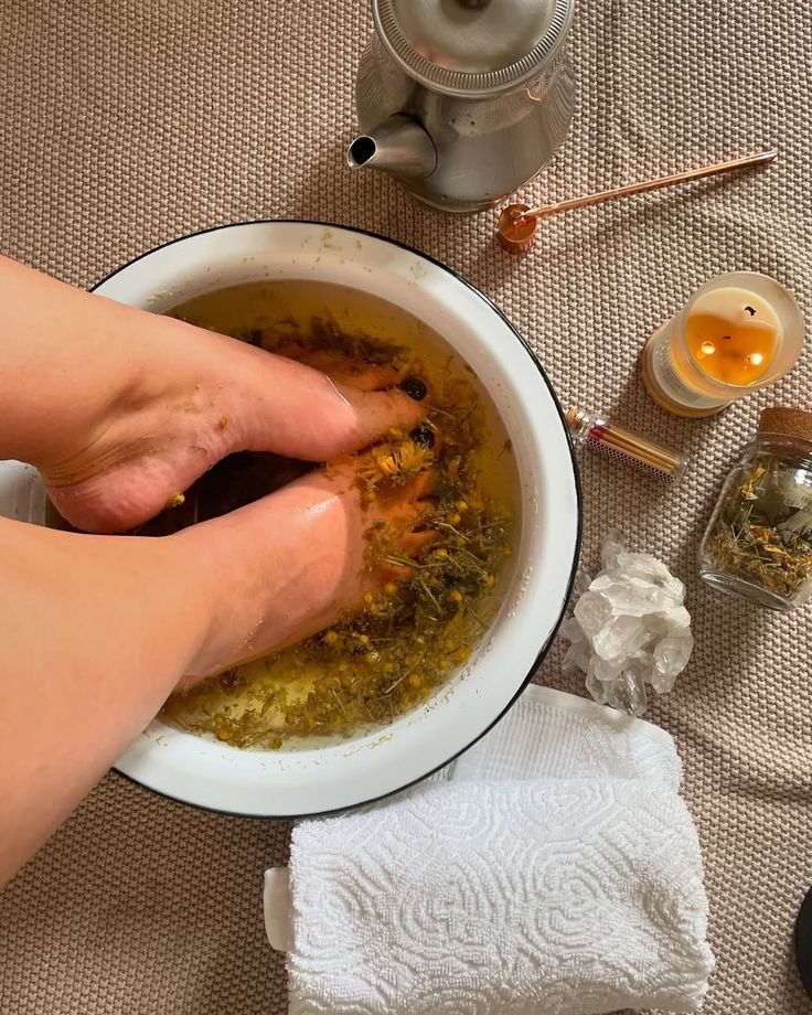 Manos remojándose en un cuenco con agua de baño de hierbas junto a una tetera, velas y una toalla.