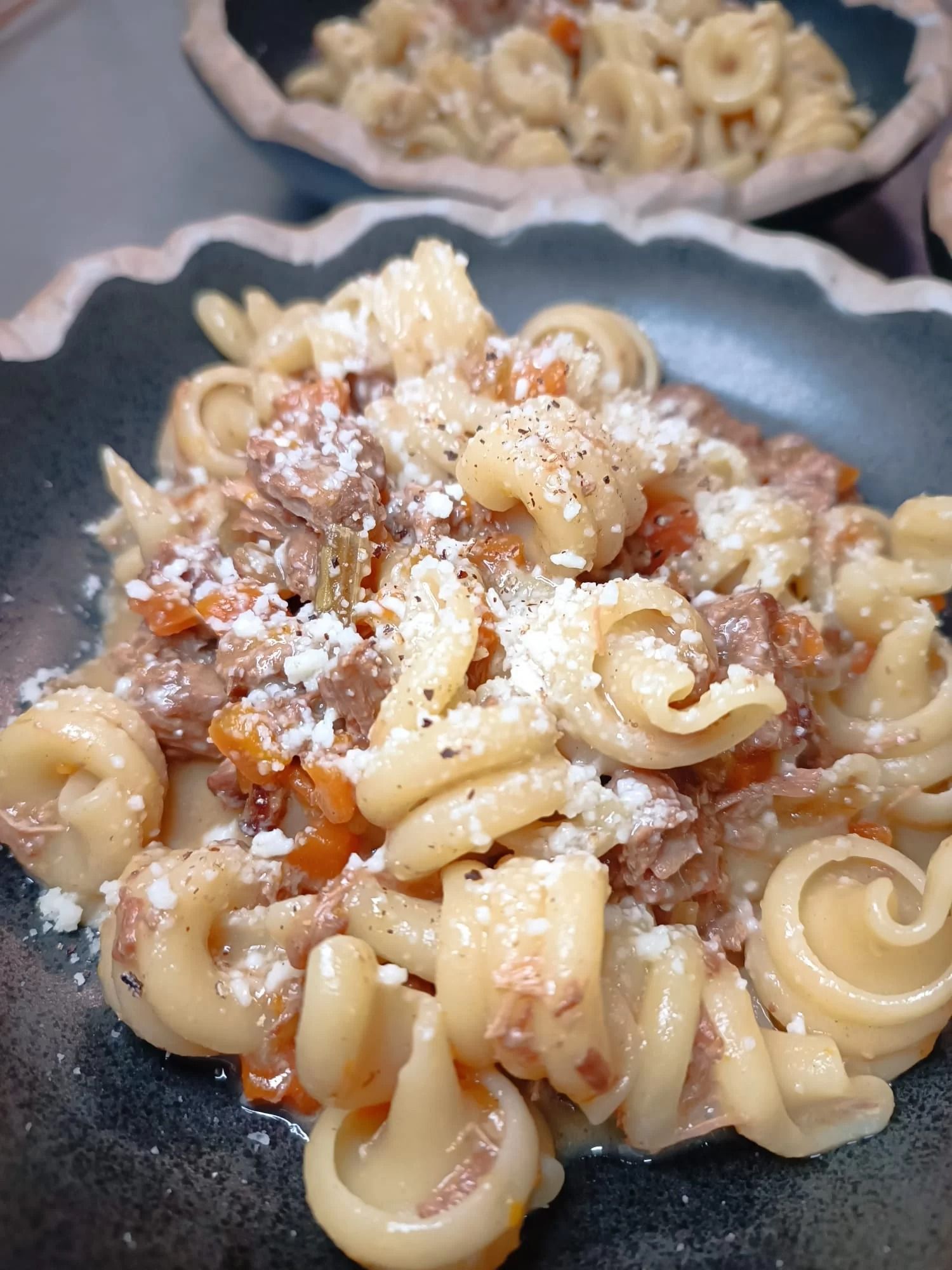 Conchiglie di pasta condite con ragù di carne e formaggio grattugiato in una ciotola scura, con altra pasta sullo sfondo.