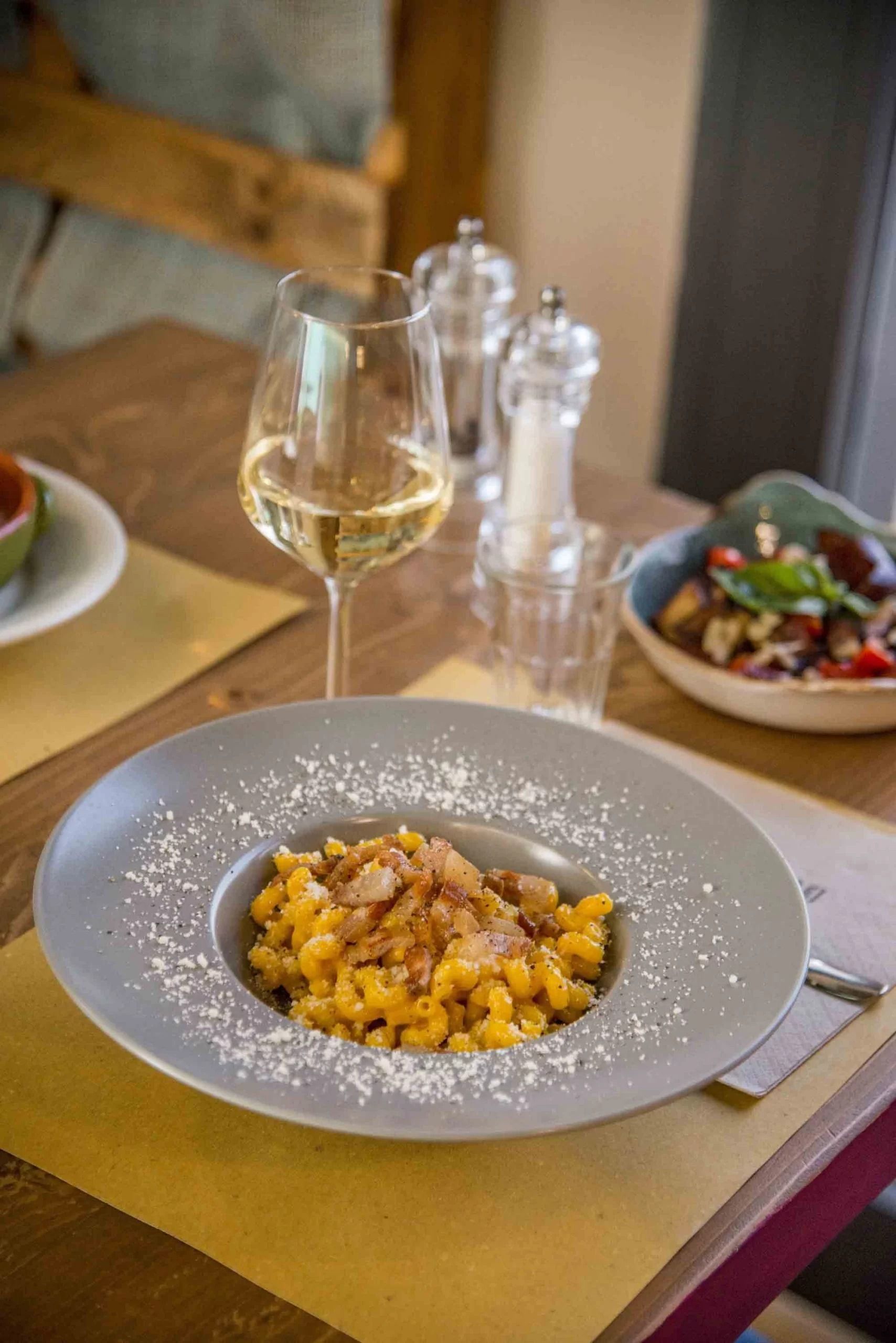 Piatto gourmet impiattato su un tavolo con un bicchiere di vino bianco e un'insalata sullo sfondo.