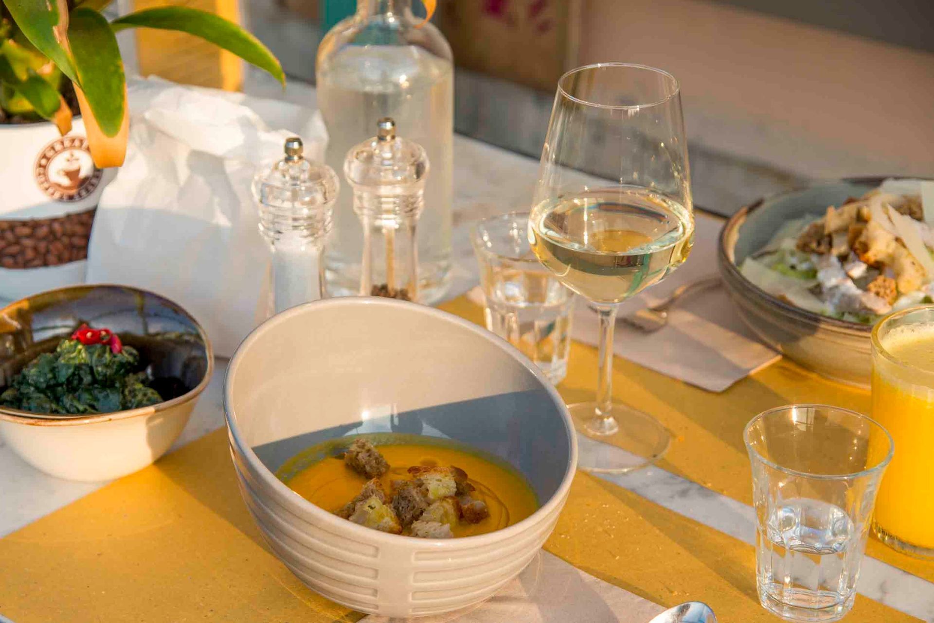 Tavolo da pranzo illuminato dal sole con ciotole di cibo, bicchieri da vino, bicchieri d'acqua e tovaglioli.