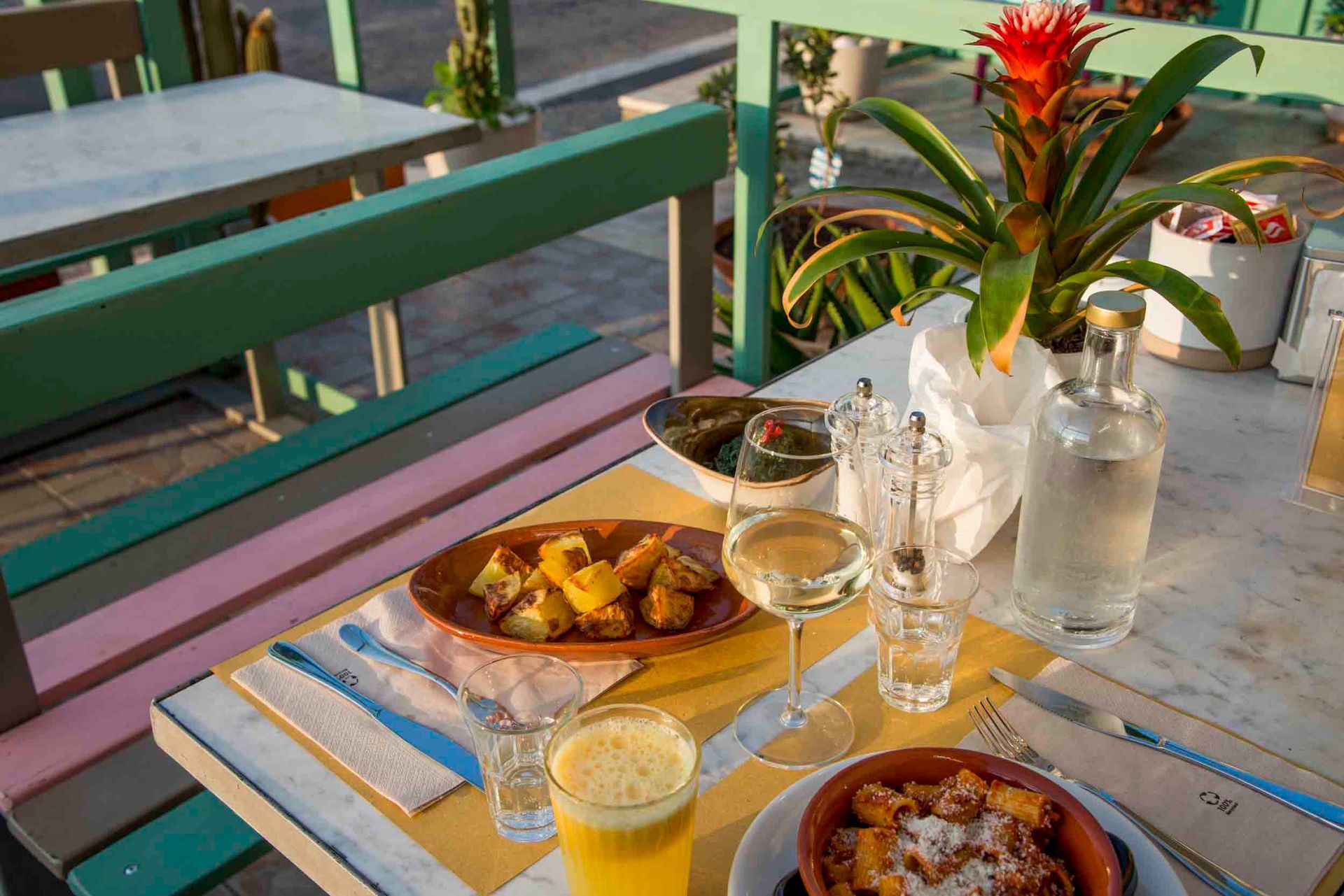 Tavolino da caffè all'aperto con ciotole di frutta, bevande e una pianta tropicale in vaso accanto a una panchina colorata.