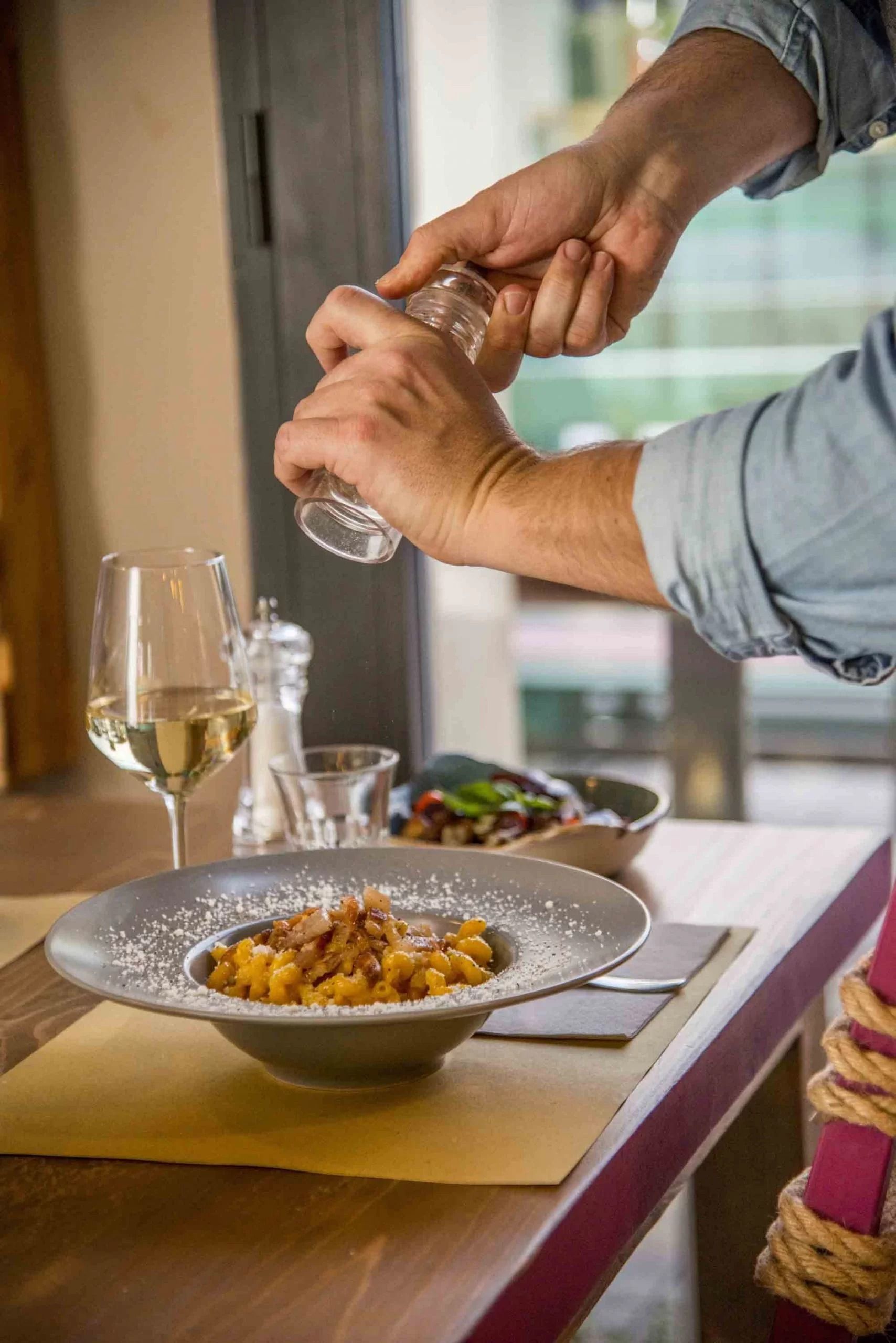 Mani che condiscono un piatto impiattato a un tavolo da pranzo con bicchieri di vino e una bottiglia nelle vicinanze.