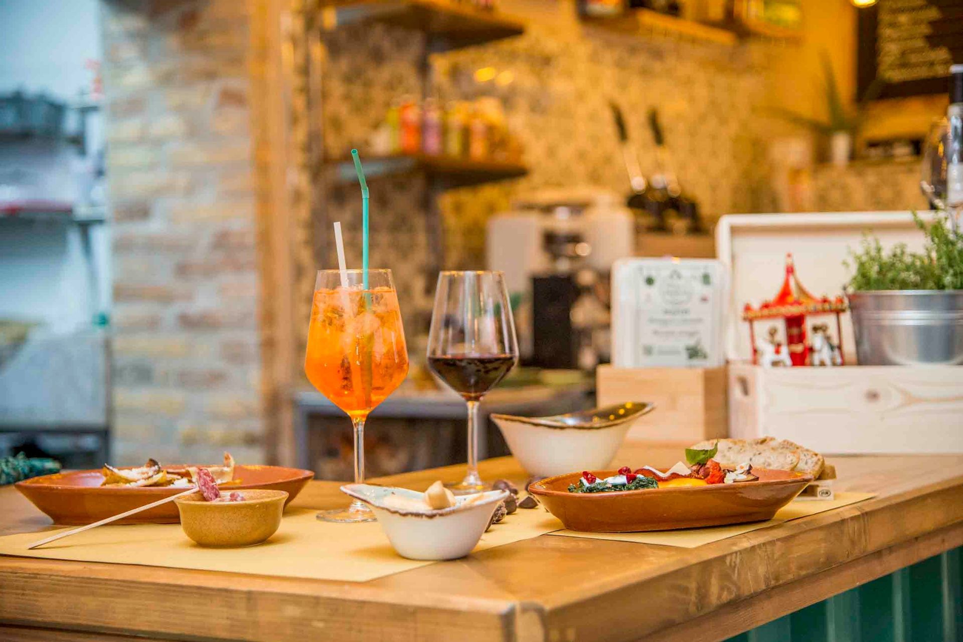 Tavolo da ristorante con antipasti, cocktail all'arancia, vino rosso e menù in un'atmosfera calda e rustica da bar.