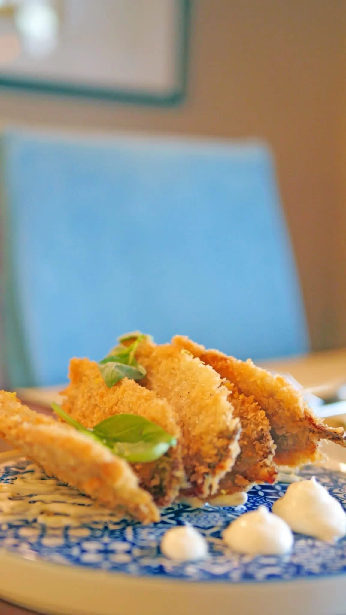 Piatto con cibo impanato e guarnizione di menta su un piatto blu e bianco, con una sedia blu sfocata sullo sfondo.