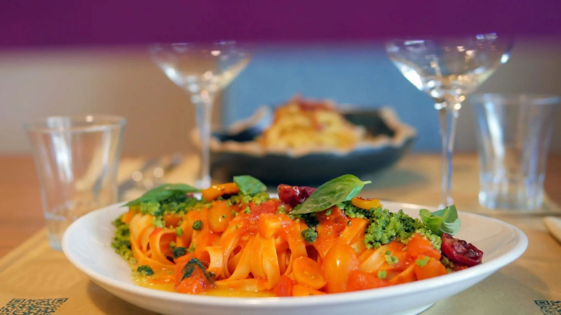 Pasta al sugo rosso e verdure su un piatto bianco, con calici di vino in una sala da pranzo leggermente sfocata.