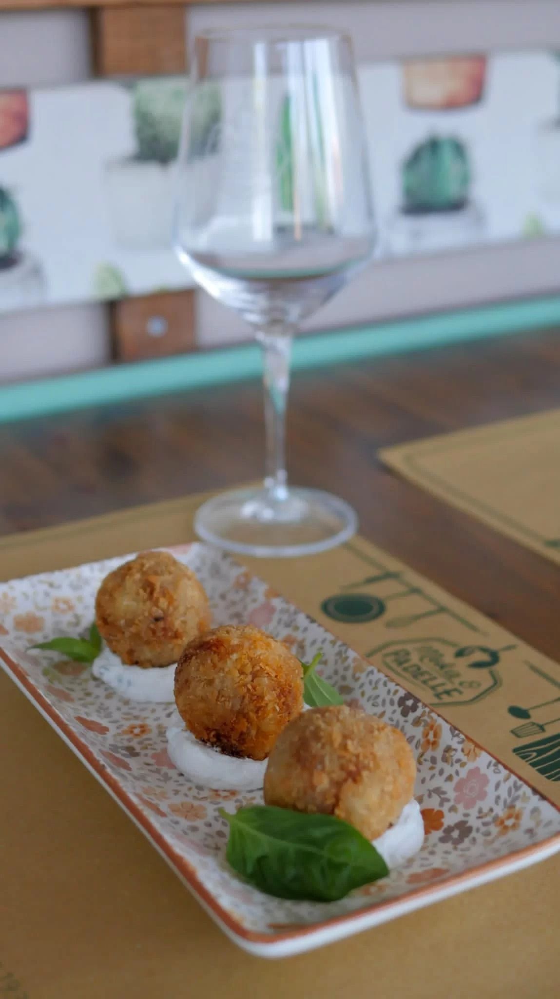 Tre crocchette fritte su un piatto decorato, accanto a una foglia di basilico e un bicchiere di vino vuoto.
