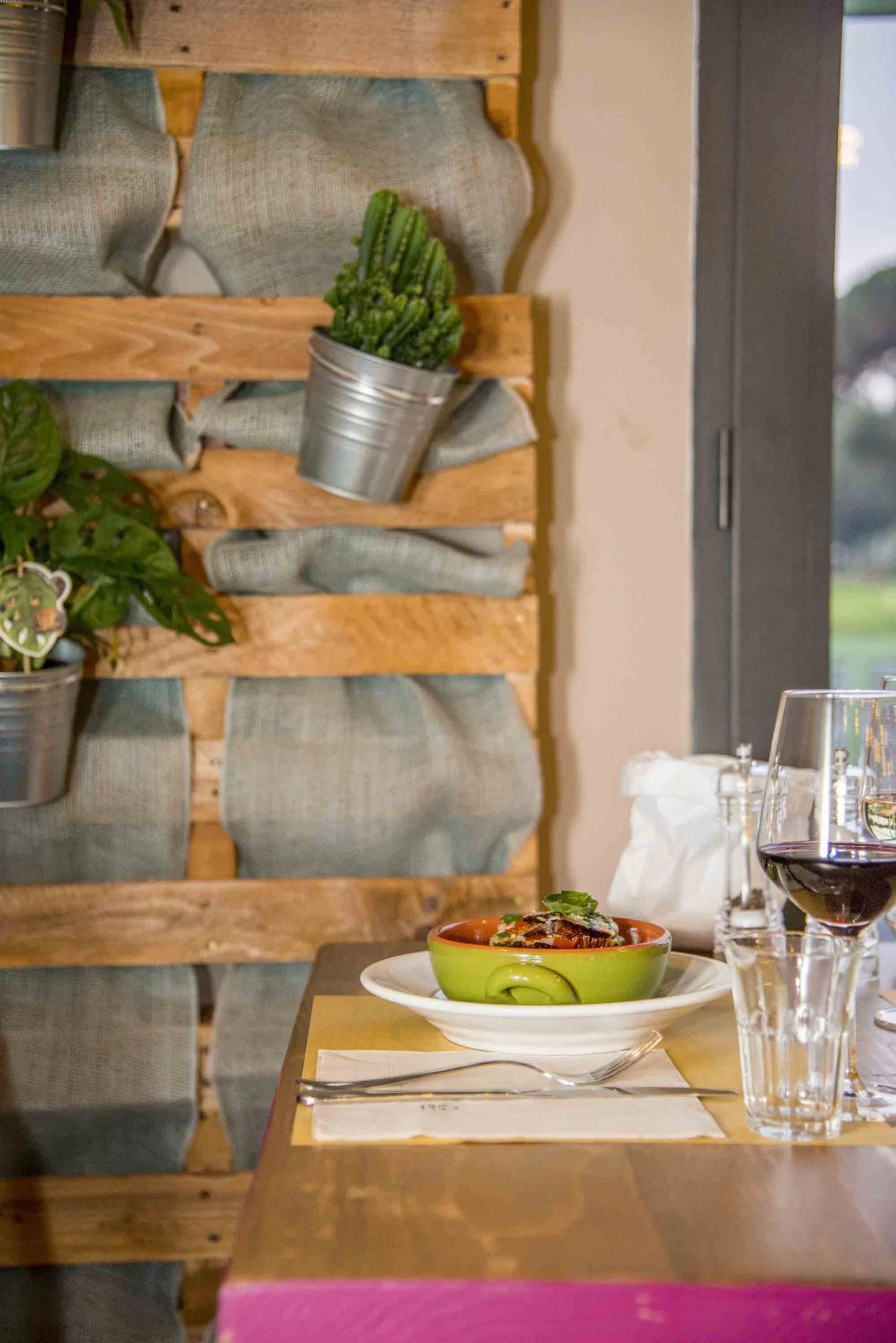 Tavolo rustico da ristorante con una ciotola di cibo, bicchieri da vino e una parete di piante sullo sfondo.