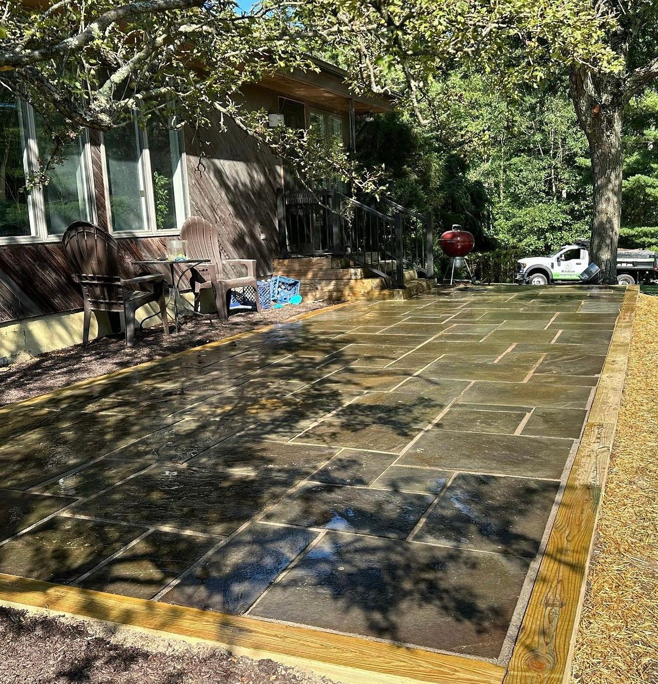 A wet, stone patio next to a house, framed by a golden border and trees. A grill, seating, and a truck are also visible.