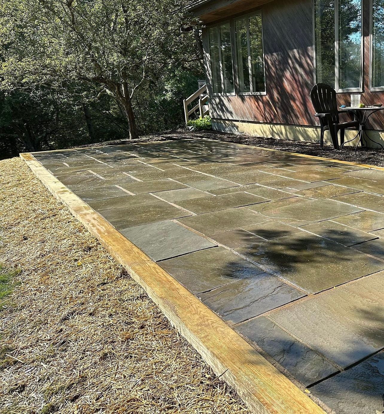 Stone patio with a wooden border, next to gravel and a house. A single chair sits on the patio.