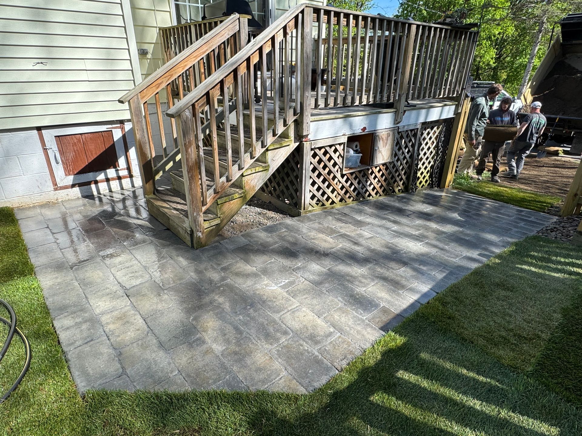 A gray paver patio beneath a wooden deck with stairs, bordered by green grass. Two people are carrying supplies near the deck.