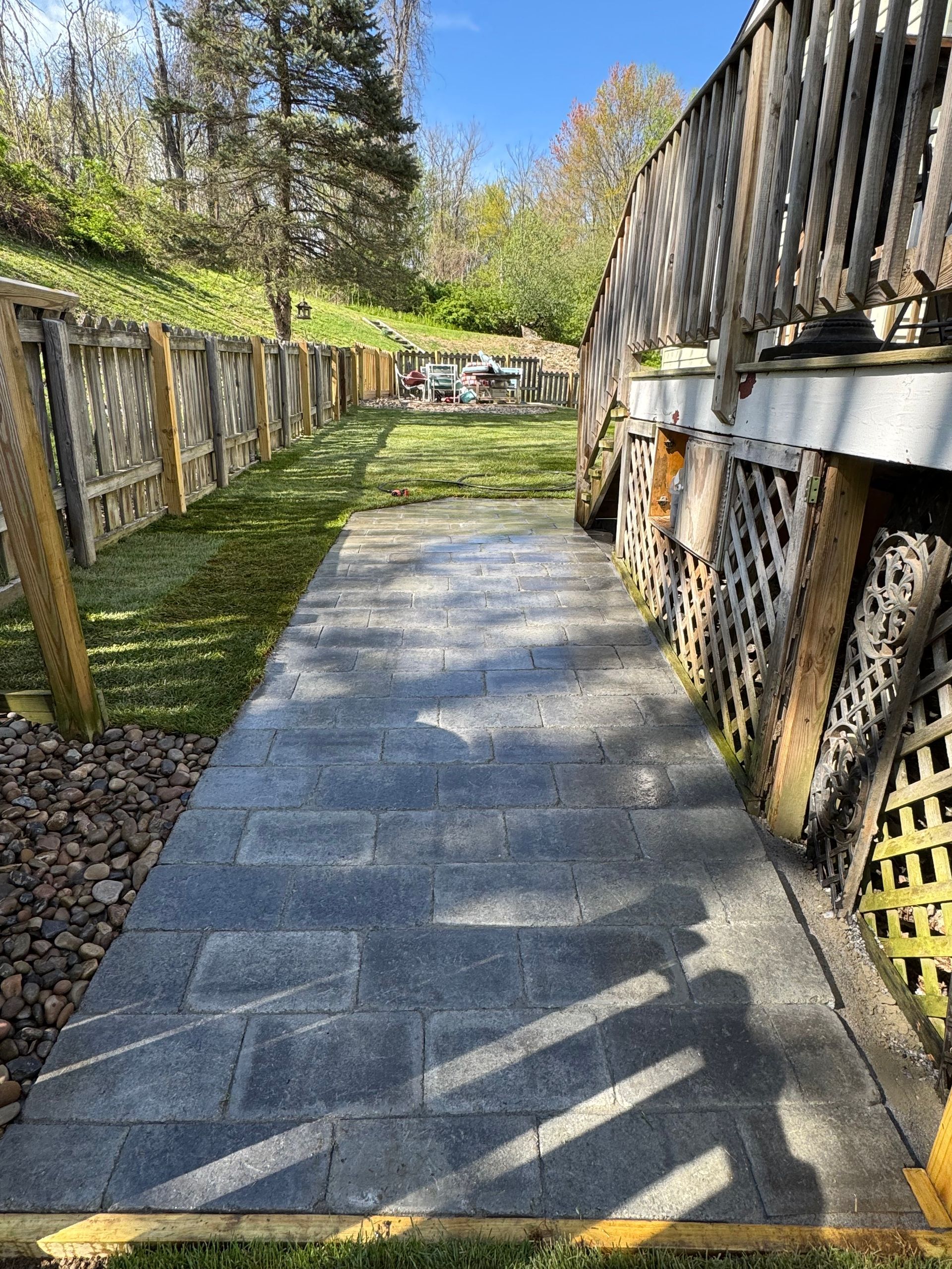 A wooden deck with stairs leading down to a gray paver patio. Green grass surrounds the patio.