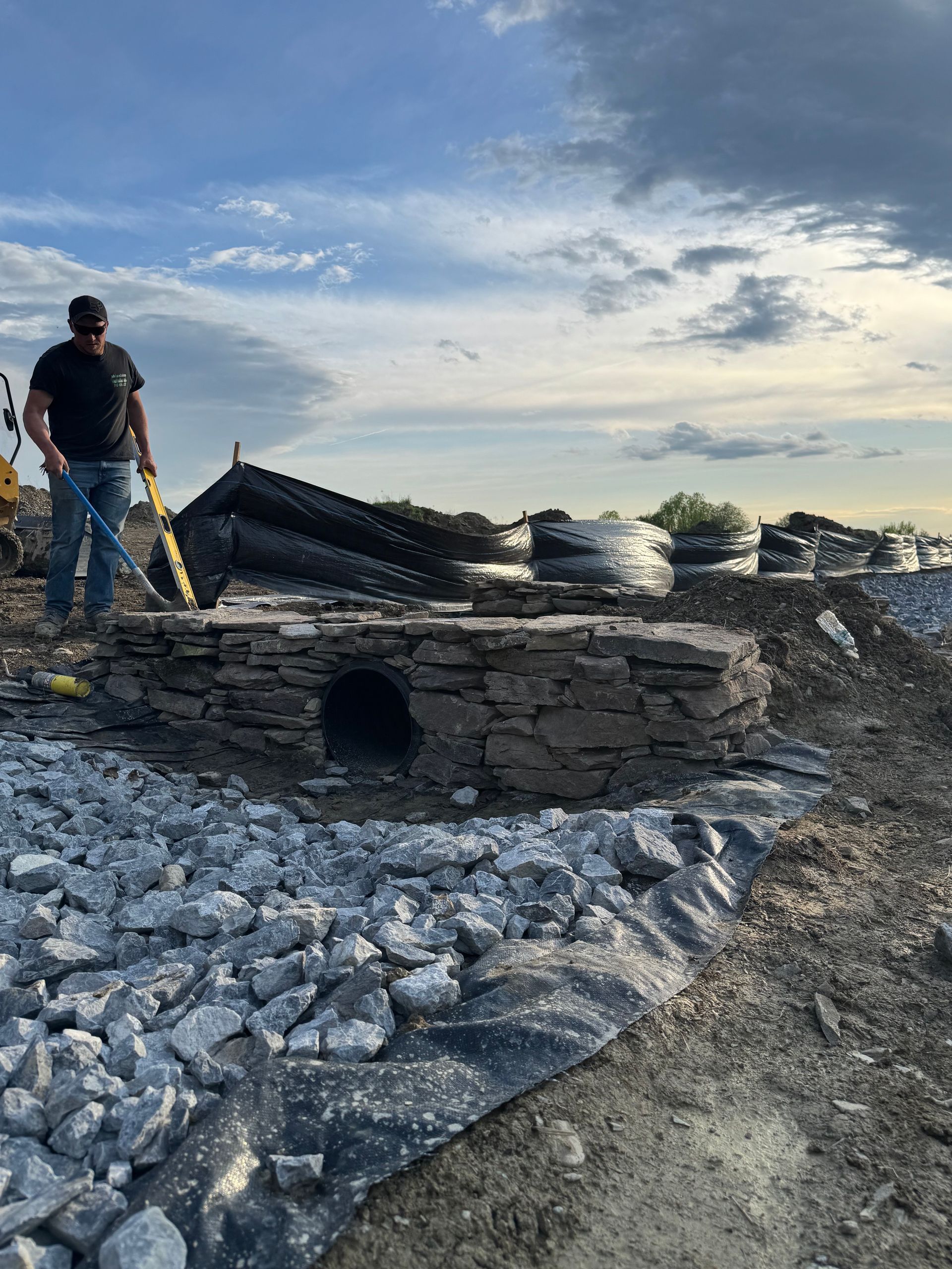 Man standing near a stone drainage structure with a round opening, surrounded by gravel and construction materials. Cloudy sky in the background.