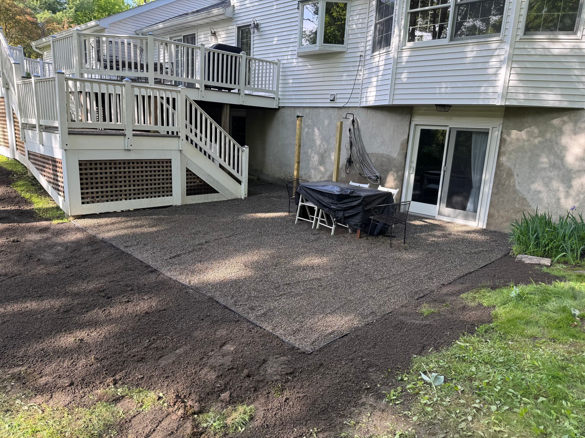 A gravel patio beside a house with a wooden deck. The patio is outlined with a darker border, and a table is on it.