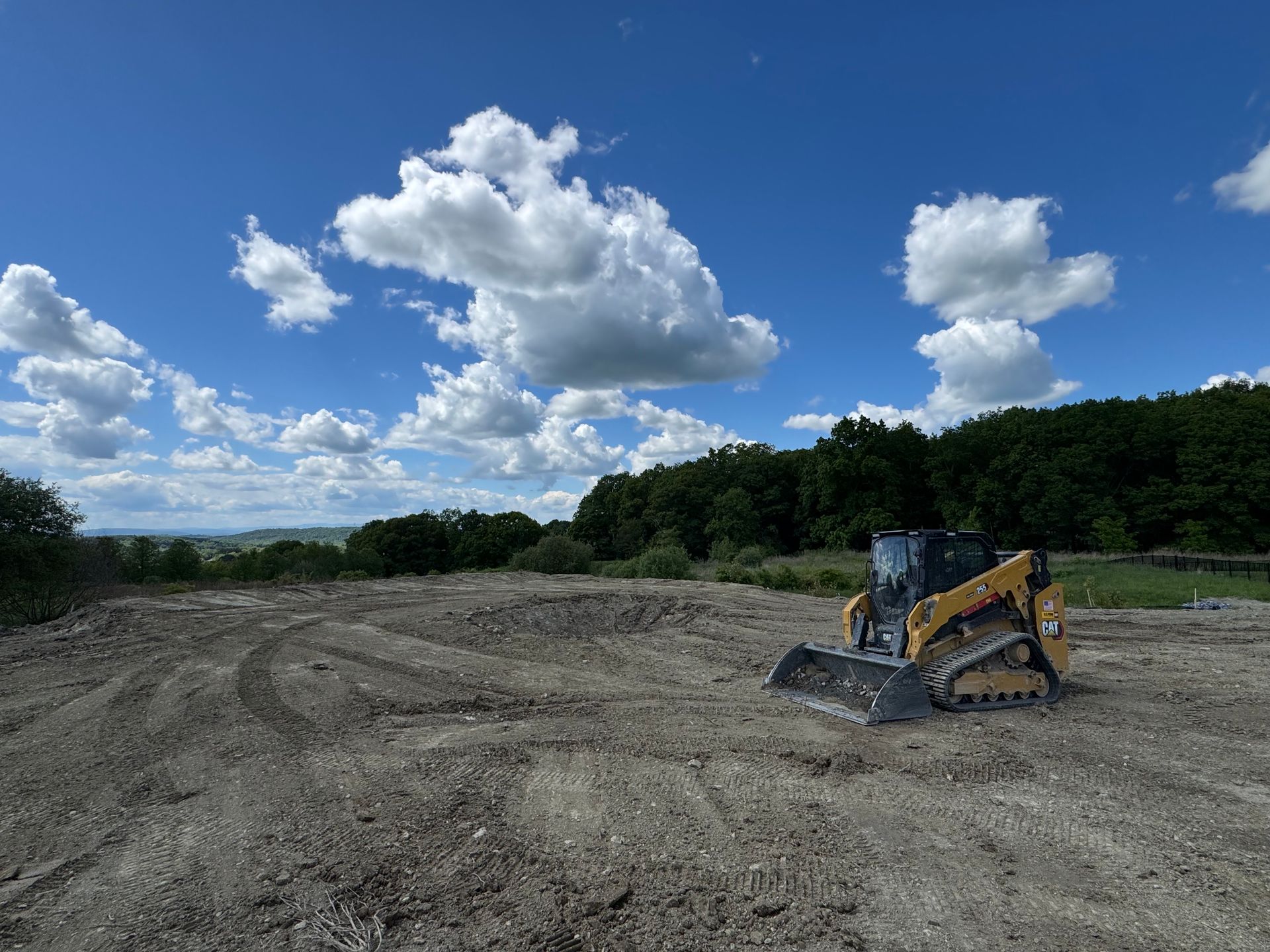 Skid steer on a dirt lot under a blue sky with puffy clouds; trees in the background.
