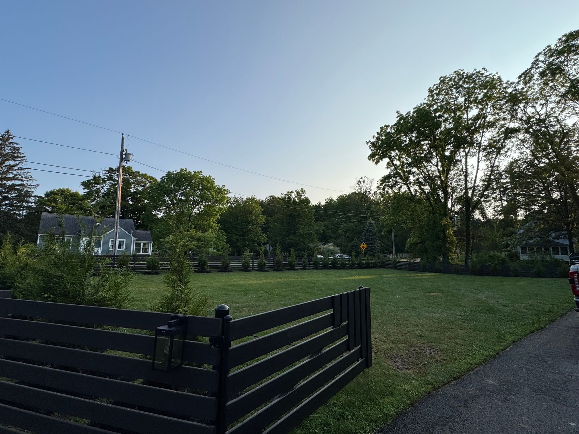 A grassy, empty lot behind a black fence, with trees and a house in the background under a blue sky.