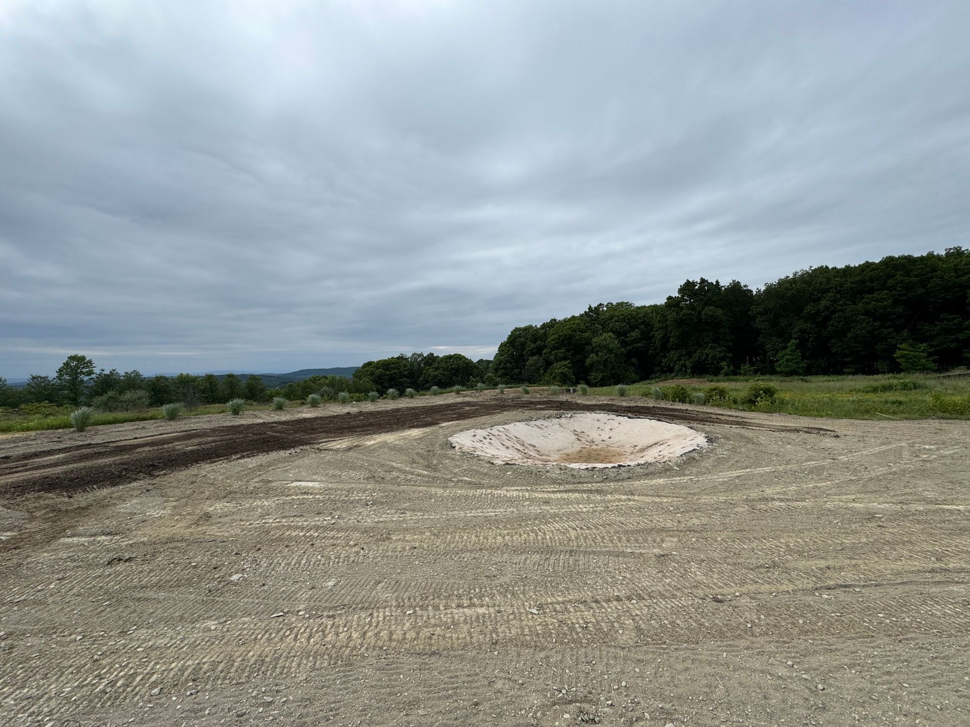 A circular, shallow depression in a gravel field, possibly a constructed feature. Overcast sky and distant treeline.